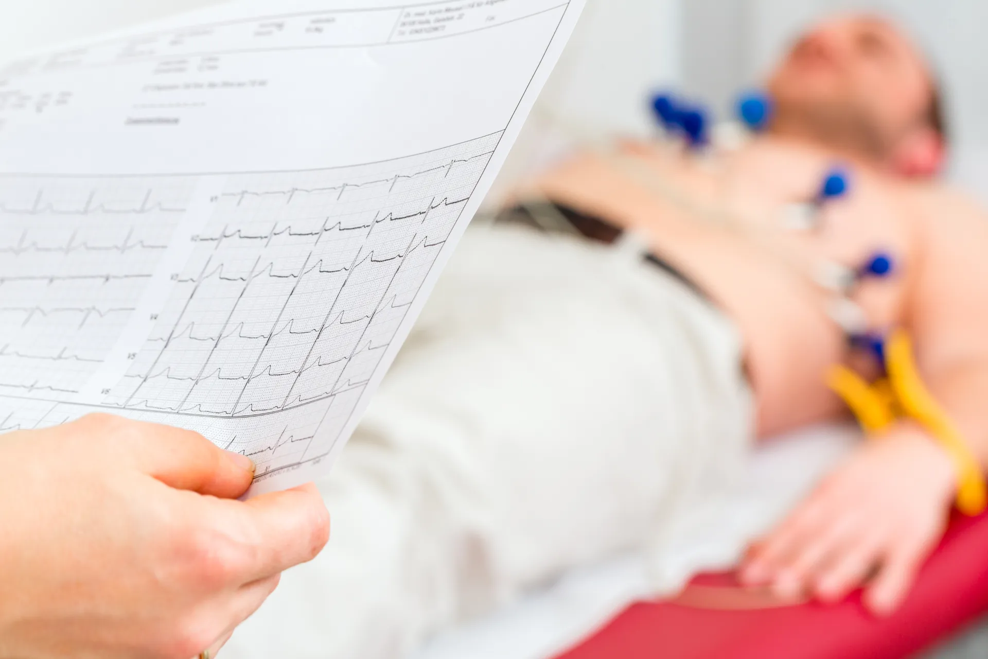Close-up of hand holding ECG report, patient undergoing electrocardiogram in background.