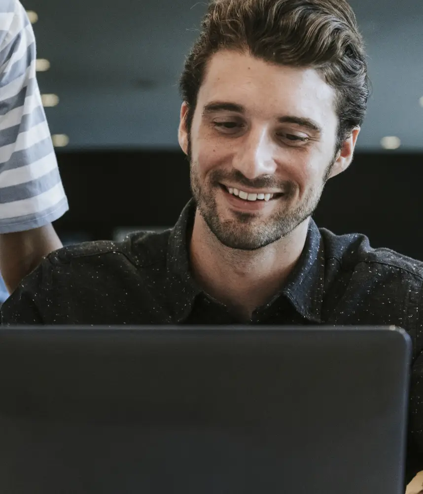 Man smiling while looking at a laptop screen, wearing a dark shirt in an indoor setting.